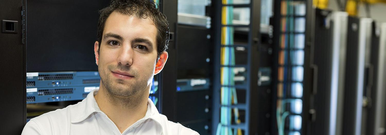 Student standing before a bank of network devices in a tower.