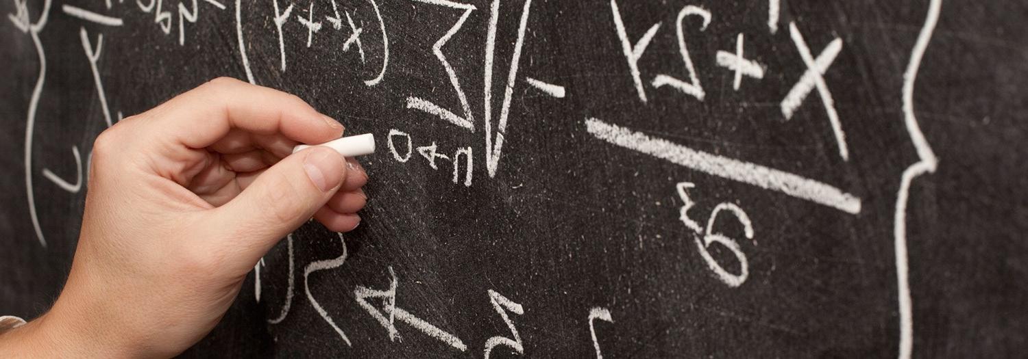 Closeup of hand writing equations with chalk on a blackboard.